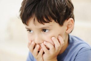 Young boy with dark hair in a blue shirt, holding his face in his hands, looking worried or thoughtful.